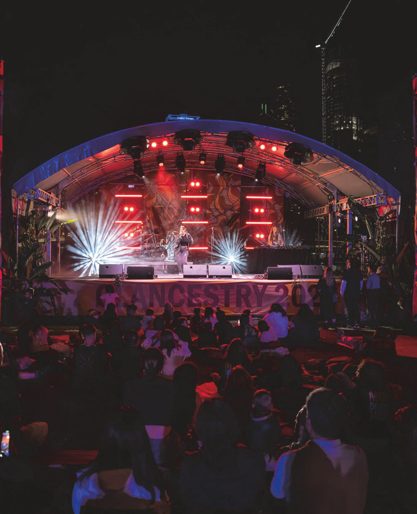 A crowd watching a lit outdoor stage at night with the Clancestry banner
